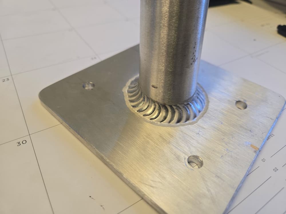 Aluminum round tube TIG welded to a base plate with a tight ring of stacked-dime weld beads.